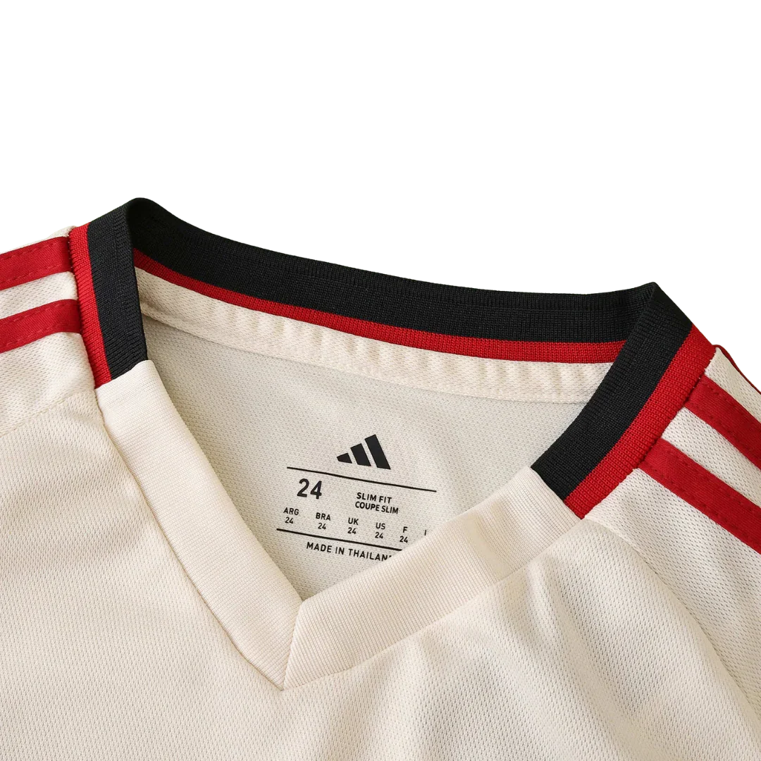 Detalhe da gola e ombro de uma camisa de futebol infantil creme com listras pretas e vermelhas. Etiqueta interna Adidas com tamanho 24 e "MADE IN THAILAND".