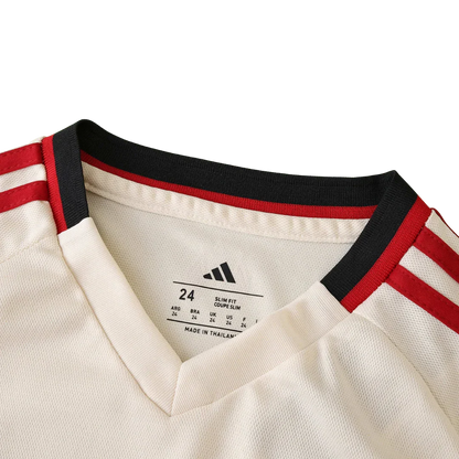 Detalhe da gola e ombro de uma camisa de futebol infantil creme com listras pretas e vermelhas. Etiqueta interna Adidas com tamanho 24 e "MADE IN THAILAND".