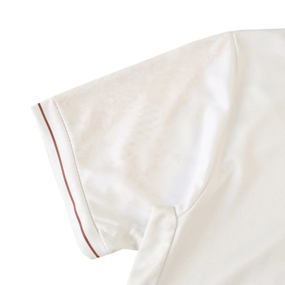 Manga de camisa infantil branca com padrão sutil e faixa marrom-avermelhada no punho. Detalhes de costura visíveis.
