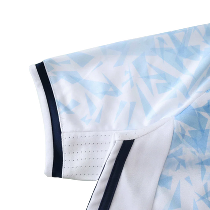 Detalhe da manga de uma camisa de futebol infantil branca com padrão geométrico azul claro, listras azul escuro e perfurações para ventilação.