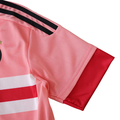 Foto aproximada da lateral de uma camisa de futebol infantil predominantemente rosa com listras pretas no ombro, listras horizontais brancas e vermelhas no corpo e punho vermelho.