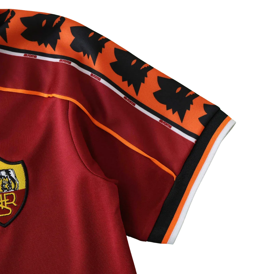 Detalhe da camisa infantil da AS Roma em tom vinho, com escudo bordado, faixas de lobos e 'ROMA', e detalhes em laranja e preto.