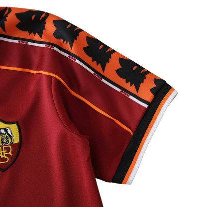 Detalhe da camisa infantil da AS Roma em tom vinho, com escudo bordado, faixas de lobos e 'ROMA', e detalhes em laranja e preto.
