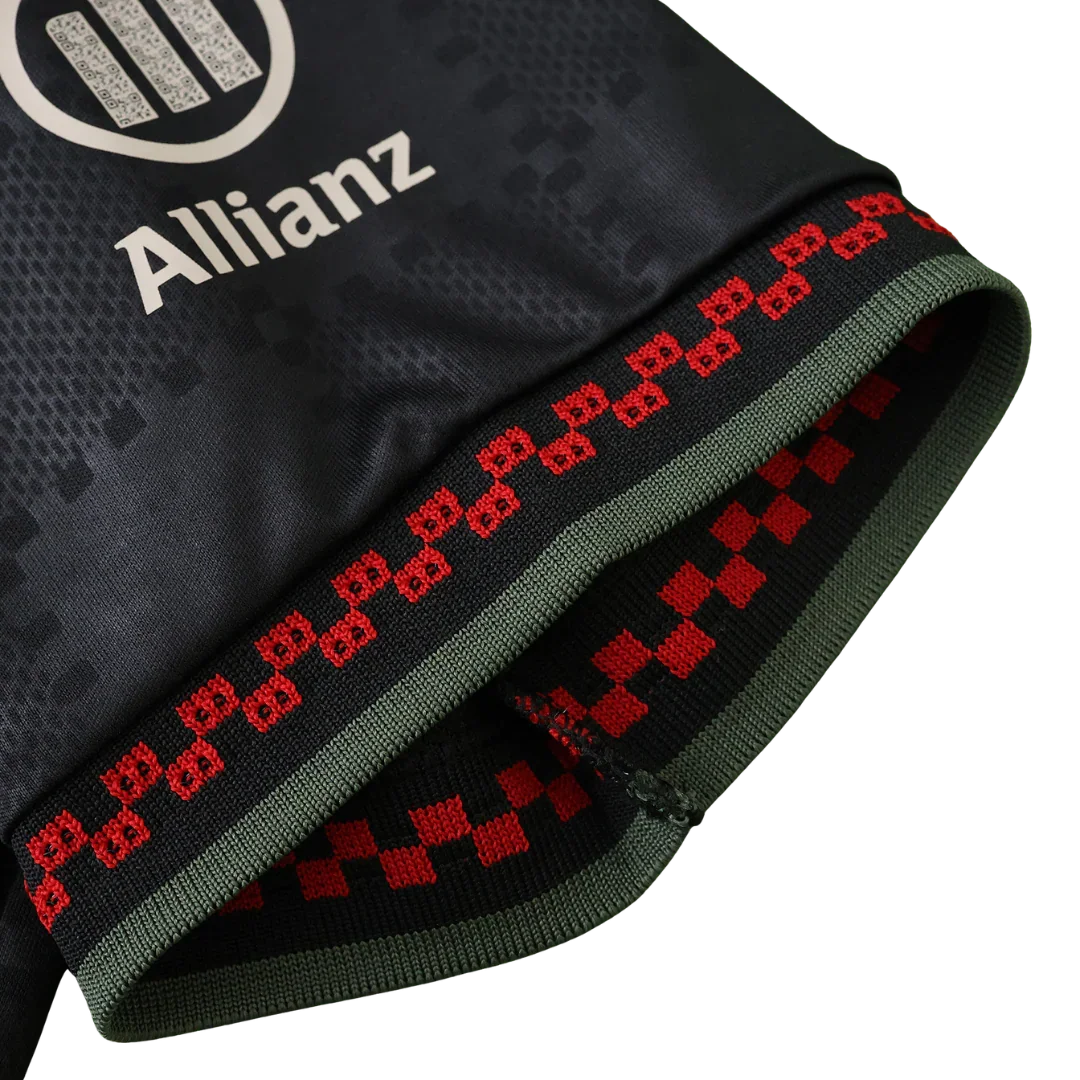 Close-up de camisa de futebol infantil escura com logo Allianz. Manga com barra preta, faixas verde-oliva, símbolos e xadrez vermelho.