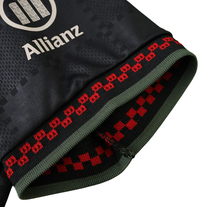 Close-up de camisa de futebol infantil escura com logo Allianz. Manga com barra preta, faixas verde-oliva, símbolos e xadrez vermelho.