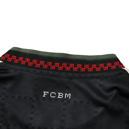 Vista traseira de camisa de futebol infantil preta com textura. Gola verde e faixa vermelha estampada. Letras FCBM brancas atrás.