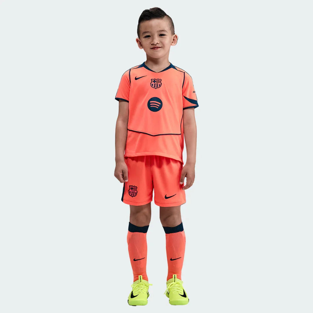 Menino com kit infantil Barcelona laranja coral vibrante, camisa, shorts e meias. Detalhes azul marinho, logos Nike e Spotify. Fundo branco.