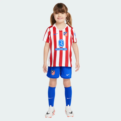 Menina sorrindo vestindo o kit infantil do Atlético de Madrid. Camisa com listras verticais vermelhas e brancas, shorts e meias azuis.
