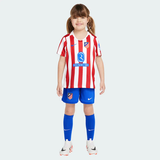 Menina sorrindo vestindo o kit infantil do Atlético de Madrid. Camisa com listras verticais vermelhas e brancas, shorts e meias azuis.