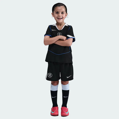 Menina sorridente veste kit de futebol infantil Chelsea III 25/26 preto com detalhes em azul e branco, e chuteiras rosa.