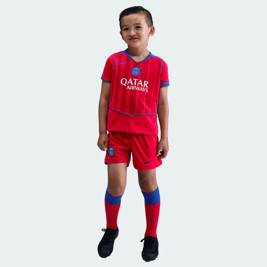 Menino sorridente veste kit de futebol infantil PSG III. Camisa e shorts vermelhos com detalhes azuis, meias vermelhas e chuteiras pretas.
