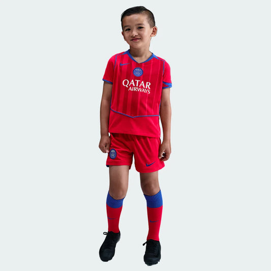 Menino sorridente veste kit de futebol infantil PSG III. Camisa e shorts vermelhos com detalhes azuis, meias vermelhas e chuteiras pretas.