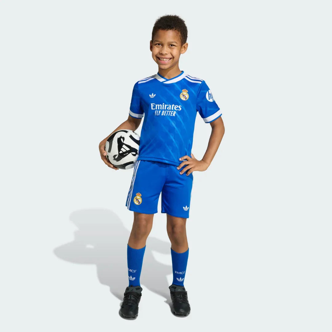 Menino sorridente em pé, vestindo kit infantil azul do Real Madrid, camisa, shorts e meias, segurando uma bola de futebol.