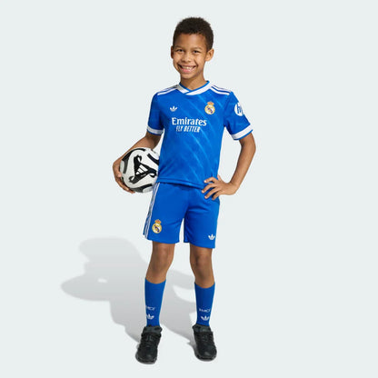 Menino sorridente em pé, vestindo kit infantil azul do Real Madrid, camisa, shorts e meias, segurando uma bola de futebol.