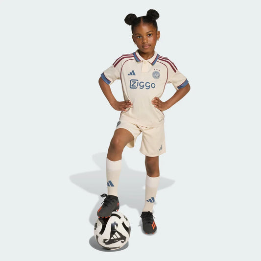 Menina veste kit infantil Ajax II 25/26 creme, com detalhes marrom e azul, shorts e meias. Pé na bola preta e branca.