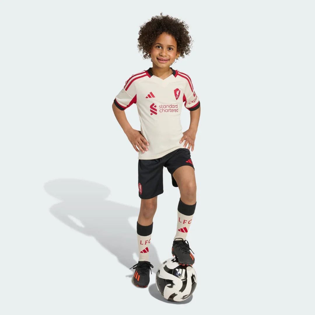 Criança sorridente em pé com kit de futebol creme e preto, bola aos pés. Camisa com escudo vermelho, patrocínio Standard Chartered.