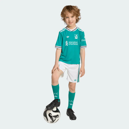 Menino sorridente com kit infantil Liverpool verde-água, shorts brancos e meias verdes-água, pé em bola de futebol.