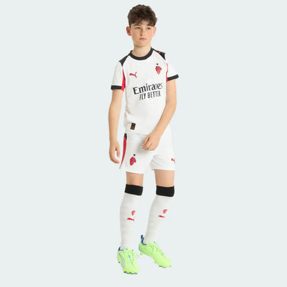 Menino em pé com kit infantil Milan II branco. Camisa, shorts e meias brancas com detalhes pretos e vermelhos. Escudo Milan, logo Puma e chuteiras verde neon.