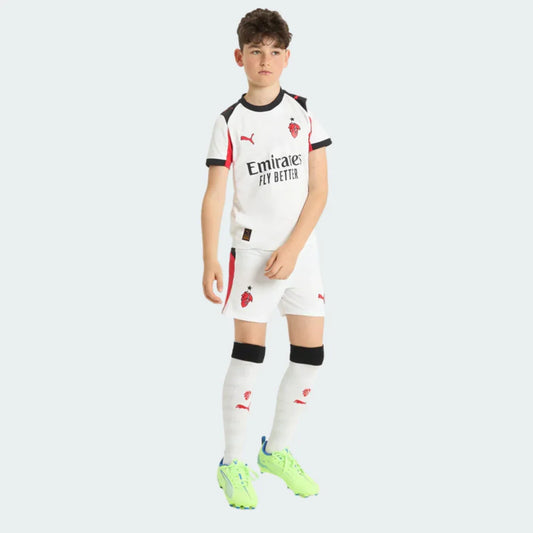 Menino em pé com kit infantil Milan II branco. Camisa, shorts e meias brancas com detalhes pretos e vermelhos. Escudo Milan, logo Puma e chuteiras verde neon.