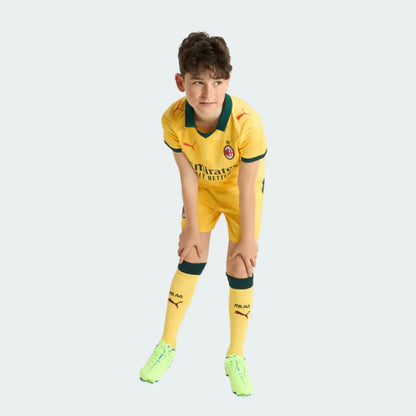 Menino usando kit infantil Milan III 25/26 amarelo com detalhes verdes. Camisa, shorts e meias combinando, chuteiras amarelas.