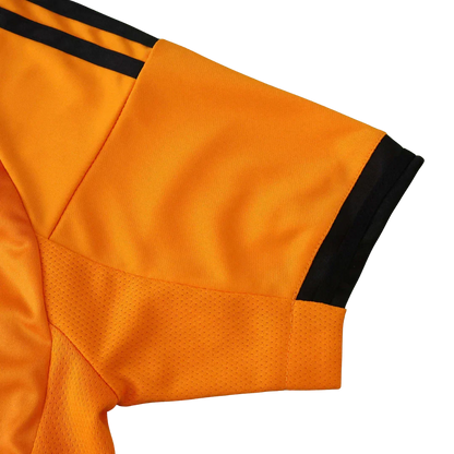 Detalhe da manga de uma camisa de futebol infantil laranja, com duas listras pretas no ombro, punho preto e painel de mesh na axila.