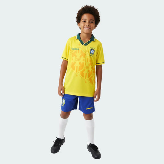 Criança sorridente veste kit de futebol retrô do Brasil: camisa amarela, shorts azuis, meias brancas e chuteiras pretas, fundo branco.
