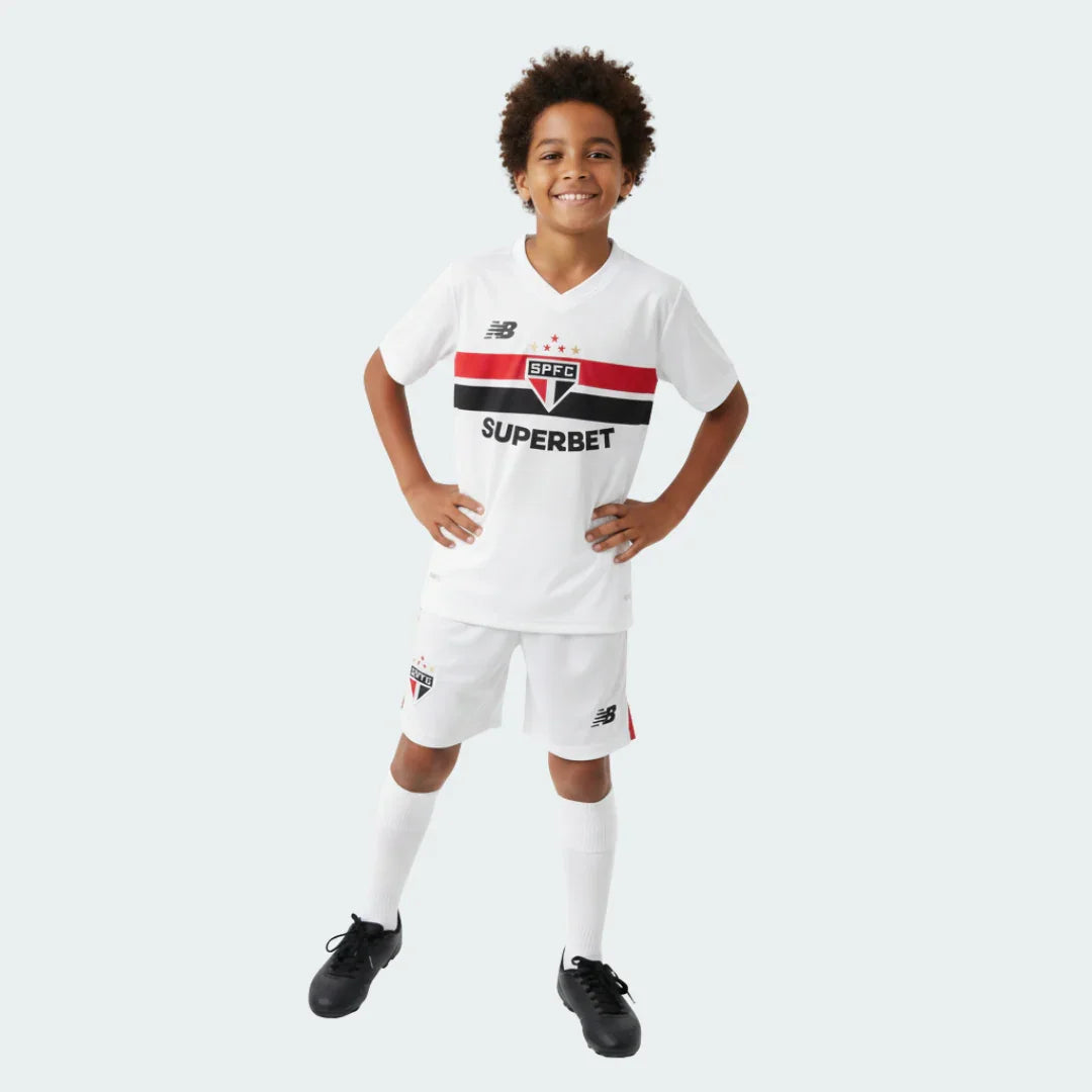 Menino sorridente vestindo kit de futebol infantil branco. Camisa com listras vermelha e preta, escudo SPFC e logos New Balance, Superbet. Shorts brancos com escudo SPFC. Chuteiras pretas.
