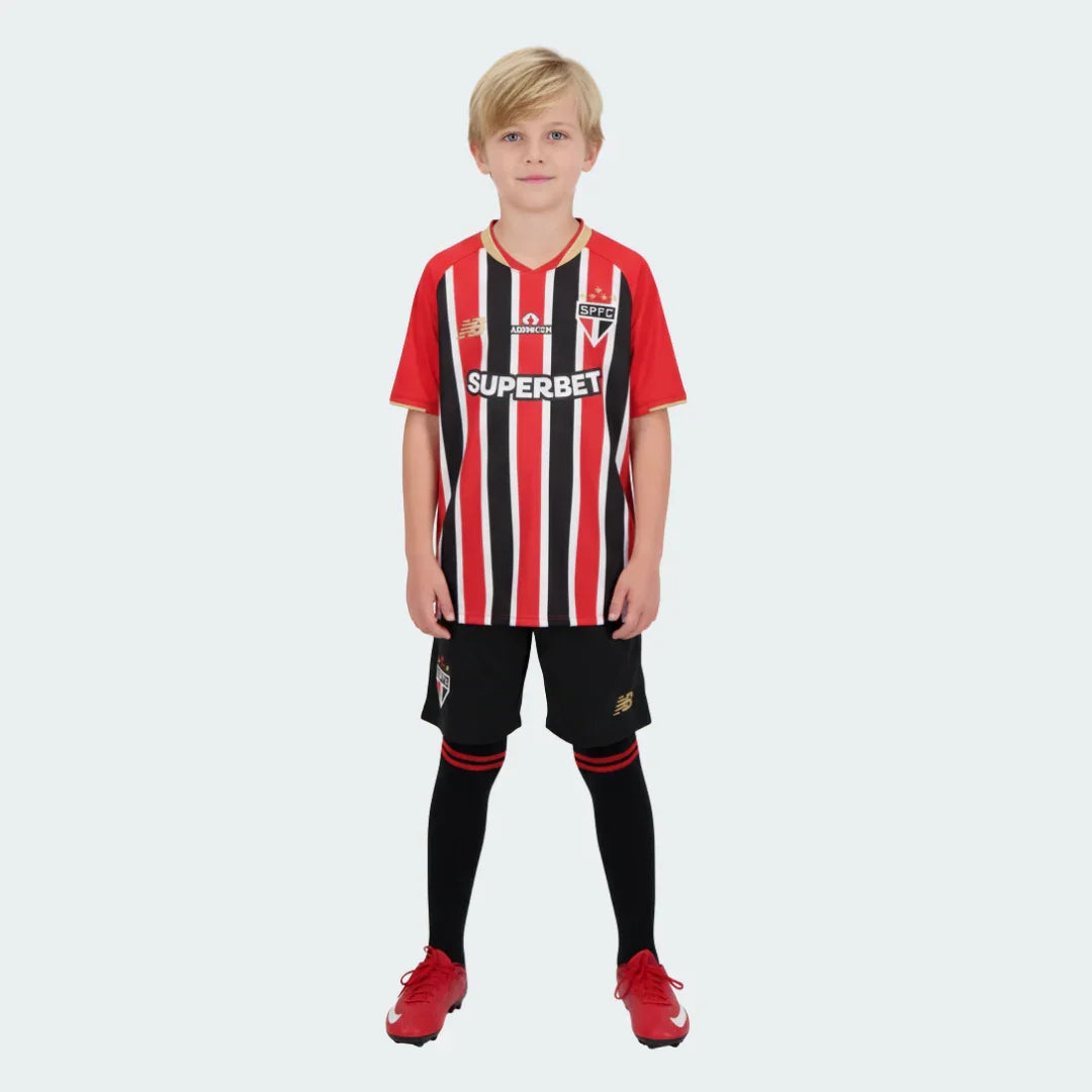 Menino em uniforme de futebol infantil do SPFC. Camisa listrada vermelha, preta e branca, shorts pretos, meias e chuteiras vermelhas.