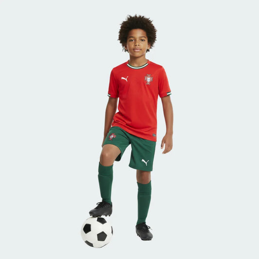 Menino com kit de futebol Portugal I 25/26, camisa vermelha, shorts e meias verdes, com bola de futebol.
