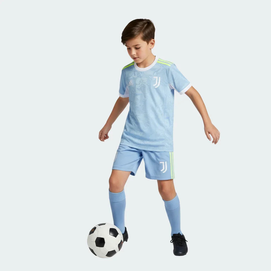 Um menino veste o kit azul claro da Juventus II 25/26 com listras amarelas, driblando uma bola de futebol.