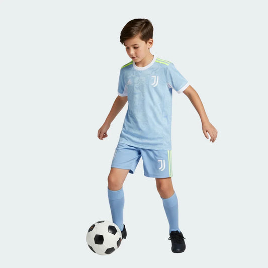 Um menino veste o kit azul claro da Juventus II 25/26 com listras amarelas, driblando uma bola de futebol.