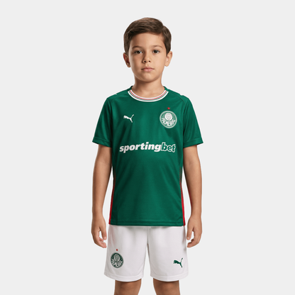 Close frontal de um menino posando seriamente, vestindo o kit infantil titular do Palmeiras na cor verde esmeralda, com gola careca branca e detalhes laterais em vermelho, acompanhado de calção branco com o escudo do clube e o logo da Puma.