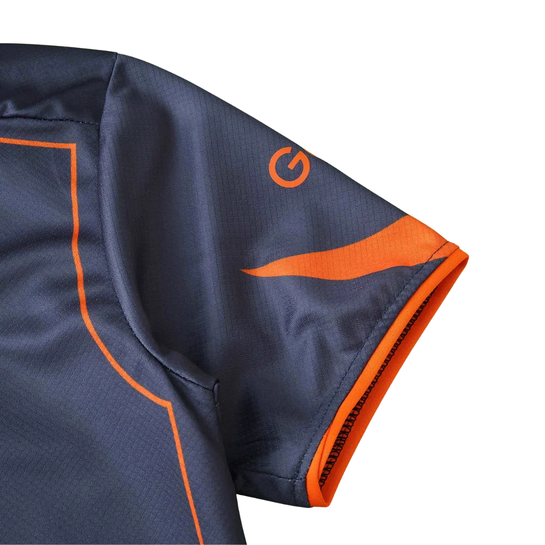 Close-up da manga de uma camisa de futebol infantil cinza escuro com detalhes laranja, incluindo um logo 'G' e barra. 