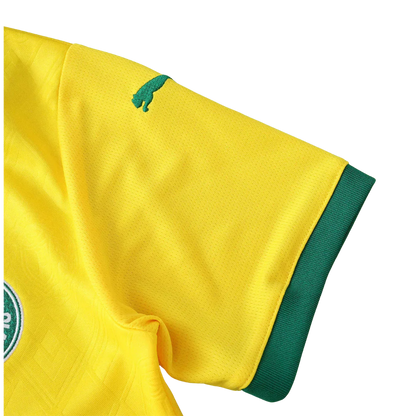 Detalhe de camisa de futebol infantil amarela com punho verde, logo verde de puma na manga e distintivo circular verde e branco.