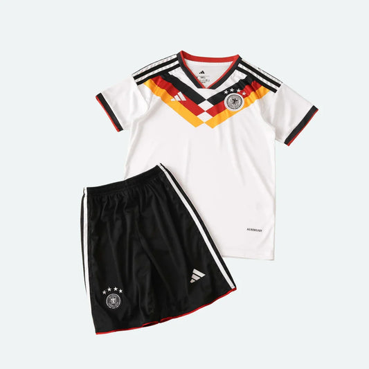 Kit infantil Alemanha 2026 com camisa branca e shorts pretos, exibindo escudos e detalhes em preto, vermelho e amarelo.