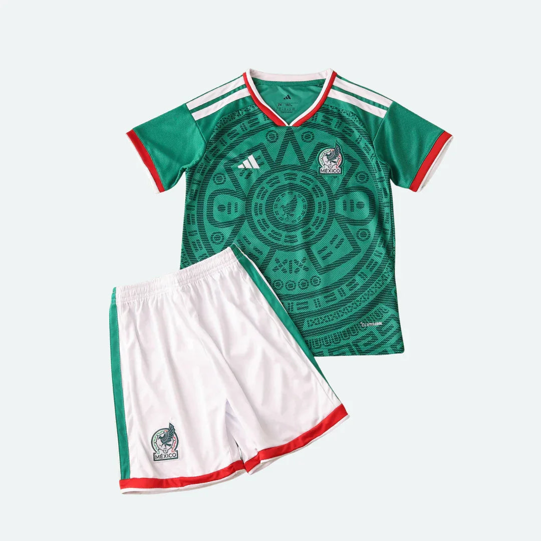 Kit infantil de futebol do México, com camisa verde escura com estampa de calendário asteca e shorts branco com listras verdes e vermelhas, sobre fundo claro.