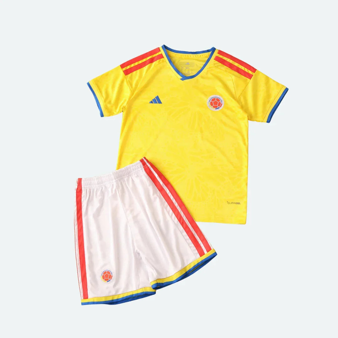 Kit infantil Colômbia I 2026 com camisa amarela, calção branco, detalhes em vermelho, azul e escudos da seleção.
