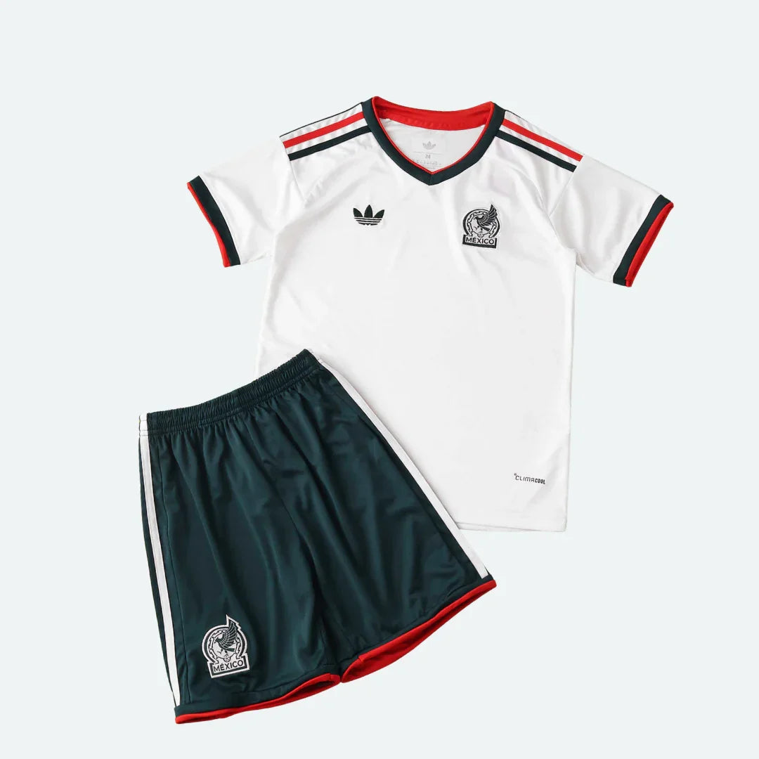 Kit infantil México II 2026 com camisa branca de gola V, detalhes em vermelho e verde escuro, shorts verde escuro com listras brancas e escudo.