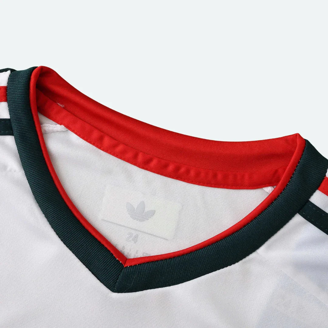 Detalhe da gola em V de uma camisa de futebol infantil branca, com acabamentos em vermelho e verde escuro.