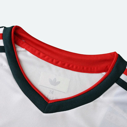 Detalhe da gola em V de uma camisa de futebol infantil branca, com acabamentos em vermelho e verde escuro.