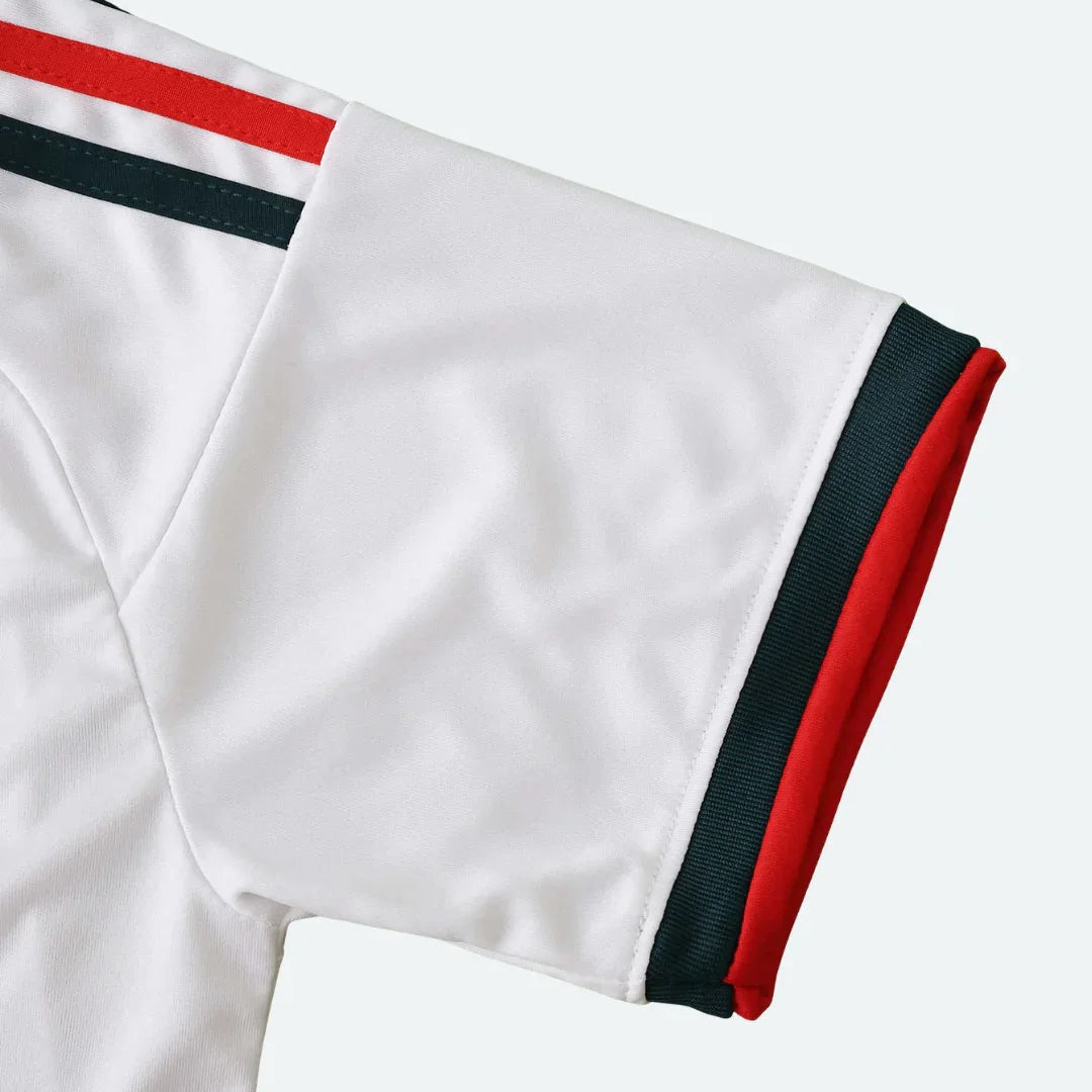 Close-up da manga de uma camisa branca. A manga tem duas listras no ombro, uma vermelha e uma verde escura, e punho com borda vermelha e faixa verde escura.