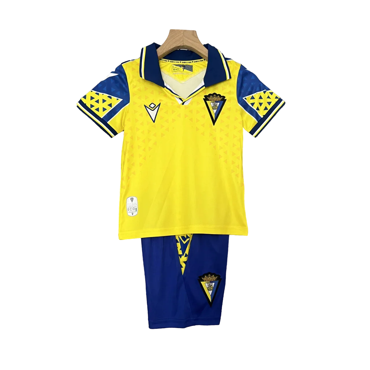 Kit infantil do Cádiz 24/25 completo com camisa amarela com detalhes azuis, escudo e shorts azuis, pendurado em fundo preto.