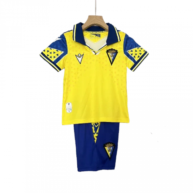 Kit infantil de futebol Cadiz I 24/25, camisa amarela com gola e detalhes azuis, shorts azuis. Em um cabide.