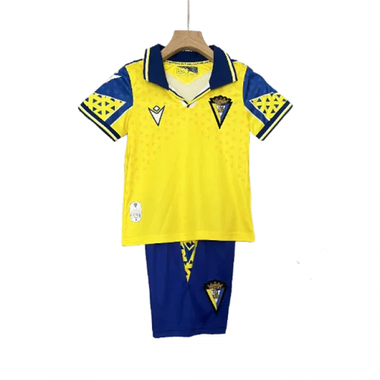 Kit infantil de futebol Cadiz I 24/25, camisa amarela com gola e detalhes azuis, shorts azuis. Em um cabide.