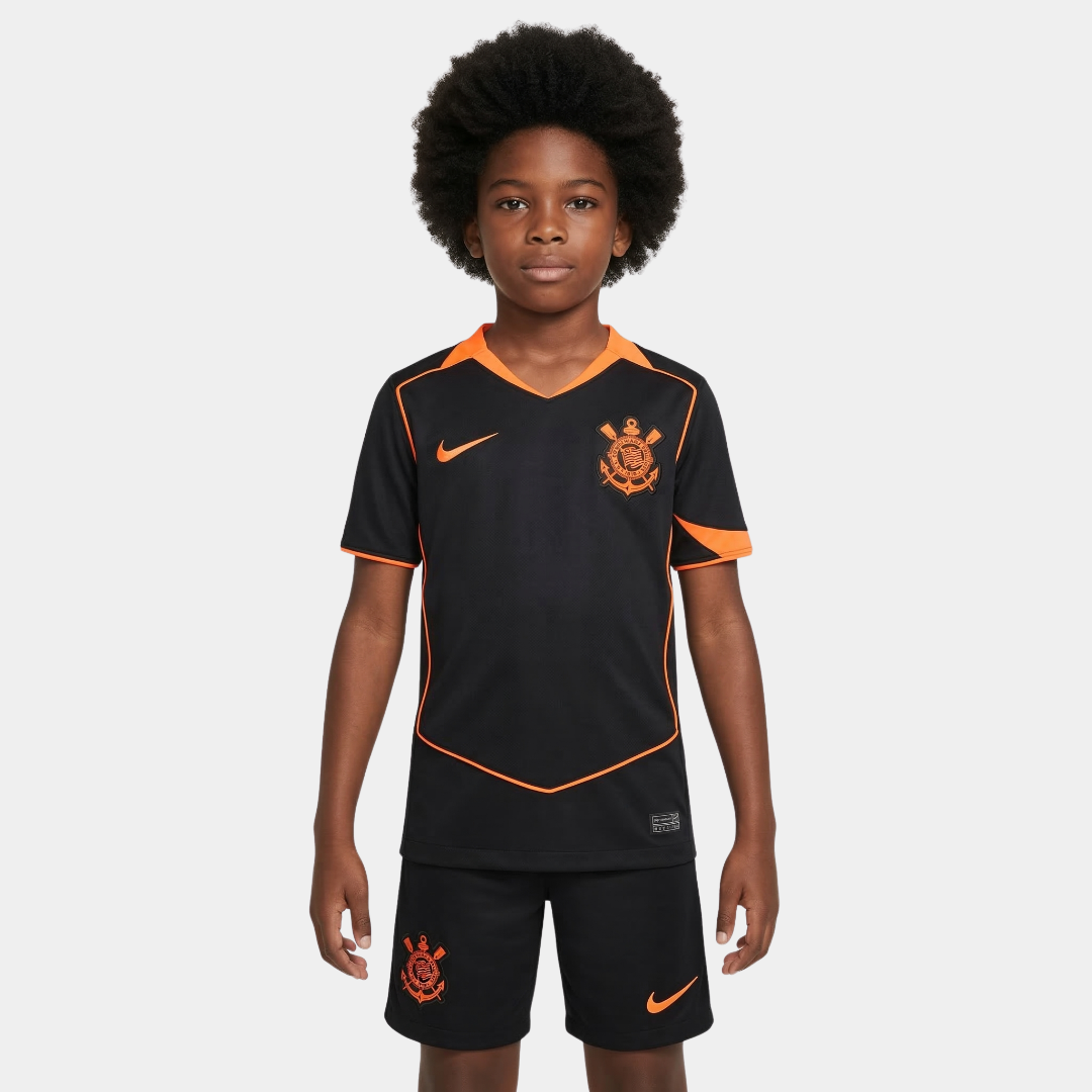 Close frontal de um menino com cabelo afro vestindo o kit infantil do Corinthians na cor preta, com detalhes em laranja vibrante na gola, ombros e laterais, acompanhado de calção preto com o escudo do clube e o logo da Nike em laranja.