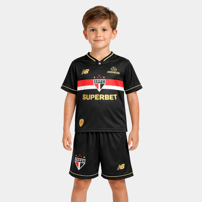 Close frontal de um menino sorridente vestindo o kit infantil do São Paulo na cor preta, apresentando uma faixa horizontal tricolor (vermelha, branca e preta) no peito com o escudo oficial centralizado, além de patrocínios e detalhes em dourado, acompanhado de calção e meiões pretos.