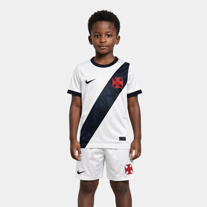 Vista frontal de um menino vestindo o kit infantil titular do Vasco da Gama na cor branca, com a icônica faixa diagonal preta atravessando o peito, ostentando a Cruz de Malta em vermelho e o logo da Nike em branco, acompanhado de calção preto com os mesmos emblemas.