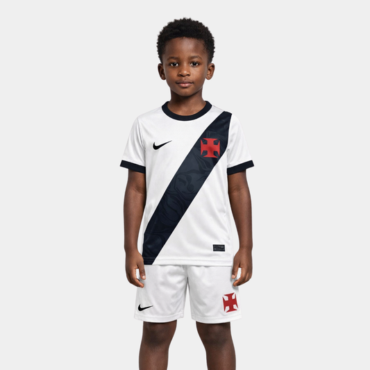 Vista frontal de um menino vestindo o kit infantil titular do Vasco da Gama na cor branca, com a icônica faixa diagonal preta atravessando o peito, ostentando a Cruz de Malta em vermelho e o logo da Nike em branco, acompanhado de calção preto com os mesmos emblemas.