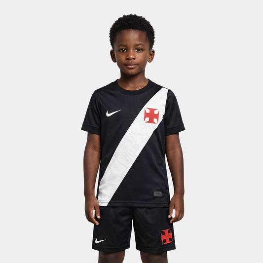 Vista frontal de um menino vestindo o kit infantil titular do Vasco da Gama na cor preta, com a icônica faixa diagonal branca atravessando o peito, ostentando a Cruz de Malta em vermelho e o logo da Nike em branco, acompanhado de calção preto com os mesmos emblemas.