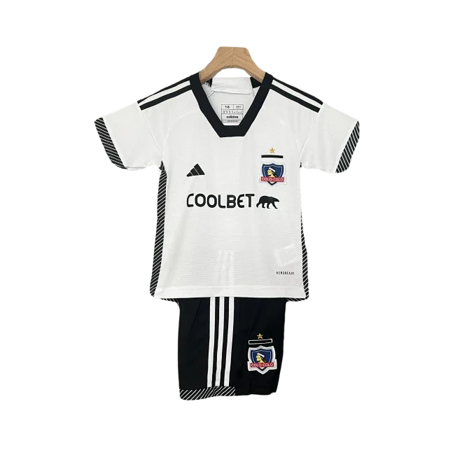 Kit infantil Colo Colo I 24/25, com camisa branca gola V preta, logos Adidas e Coolbet pretos, escudo. Shorts pretos com listras brancas.
